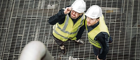 Zwei Bauarbeiter in Schutzwesten und Helmen blicken von oben auf Metallgitter herab und lächeln.