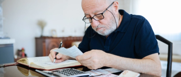 Ein älterer Mann mit Brille sitzt an einem Tisch und überprüft konzentriert Rechnungen und Belege