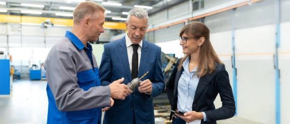 Eine Unternehmerin und ein Unternehmer stehen in einer Werkhalle und besprechen ein Projekt im Vordergrund laeuft ein Mitarbeiter an der Gruppe vorbei