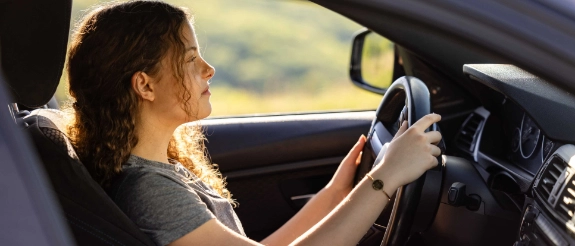 Frau mit Zopf hält Lenkrad und schaut auf Smartphone während sie Auto fährt, grüne Landschaft im Hintergrund