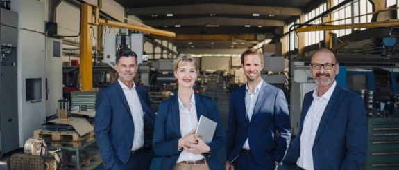 Vier Geschäftsmänner in dunklen Anzügen stehen in einer Industriehalle mit Maschinen und Produktionsanlagen im Hintergrund.