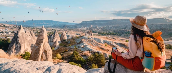 Frau mit Rucksack sitzt auf Felsen und blickt auf Kappadokien-Landschaft mit Feenkaminen und Heißluftballons