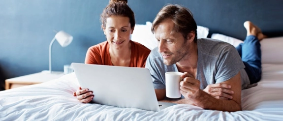 Paar sitzt im Bett vor Laptop, Frau in orangem Shirt lächelt, Mann in blauem Shirt hält Kaffeetasse, blaue Wand im Hintergrund