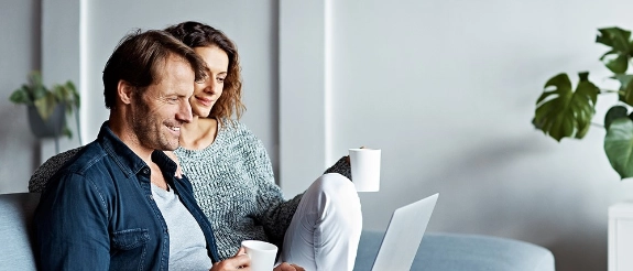 Paar sitzt zusammen und schaut auf Laptop, hält Kaffeetassen, helles Büro-Interieur