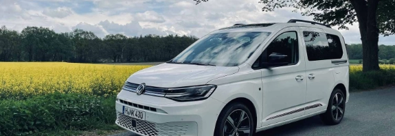 Weißer VW ID.Buzz auf Landstraße vor gelbem Rapsfeld und grünem Wald bei Tageslicht