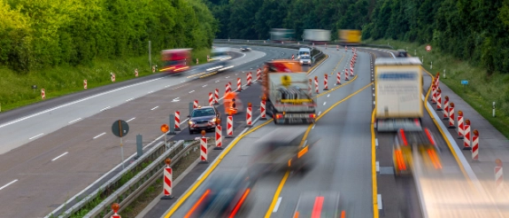 Luftaufnahme einer Autobahn mit mehrspurigen Fahrspuren, bunten Markierungen und Lastkraftwagen, umgeben von grünen Wäldern