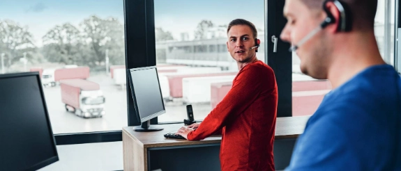 Zwei Männer im Büro, einer in rotem Hemd am Schreibtisch, anderer in blauem Hemd mit Headset, arbeiten am Computer.