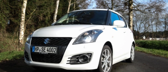 Weißer Suzuki Swift auf Parkplatz mit Birken und kahlen Bäumen im Hintergrund bei Tageslicht