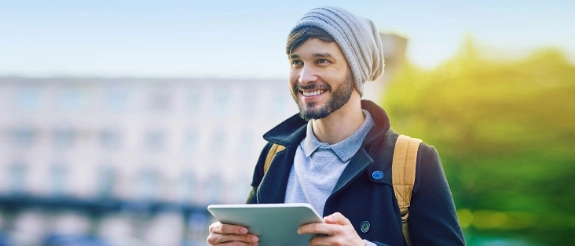 Lächelnder Mann mit Bart und Beanie hält Tablet in der Hand, trägt Jacke und Rucksack draußen
