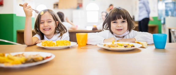 Zwei Kinder sitzen am Tisch mit Tellern und Getränken, im Hintergrund weitere Personen in einer hellen Schulkantine oder Kindertagesstätte.
