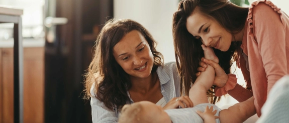 Eine junge Mutter betrachtet liebevoll gemeinsam mit einer Freundin ihr Baby. Die Freundin ist die Patentante des Babys. Sie denken über das Sparen für Patenkinder nach.
