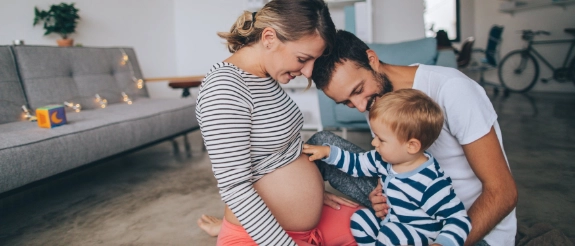 Kleiner Junge sitzt auf dem Schoss seines Vaters und fasst den schwangeren Bauch seiner Mutter an.