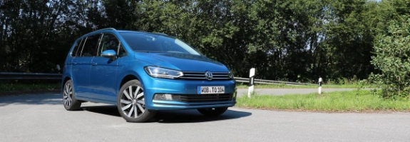 Blauer VW Golf parkt auf grauem Asphalt vor grünen Bäumen und blauem Himmel