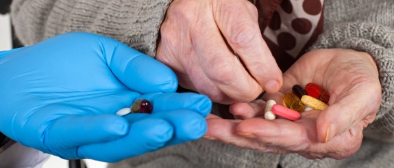 Senior mit Tabletten in der Hand nimmt noch mehr Tabletten aus der Hand einer Pflegeperson mit Handschuhen an.