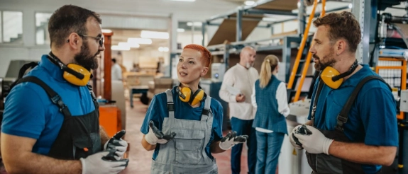 Handwerker und Auszubildende in blauer Arbeitskleidung in einer Werkstatt, diskutieren und lachen zusammen.