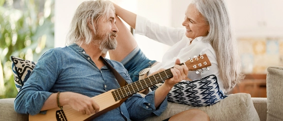 Älterer Mann spielt Gitarre, während ältere Frau neben ihm auf der Couch sitzt und zuhört