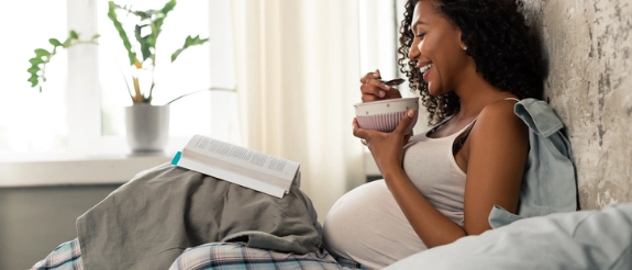 Schwangere Frau sitzt im Bett, hält eine Schüssel und isst, während sie ein Buch liest. Helles Schlafzimmer mit Pflanze.