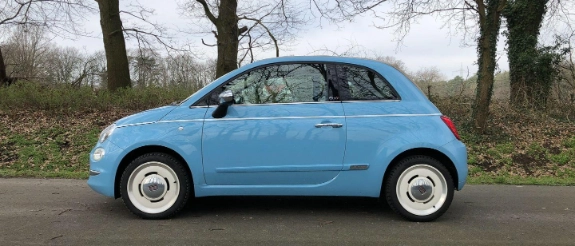 Blauer Fiat 500 Kleinwagen steht auf grauer Asphaltstraße, unbewaldete Bäume im Hintergrund