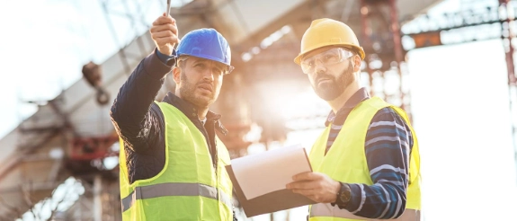 Zwei Bauarbeiter in Schutzwesten und Helmen besprechen ein Projekt auf einer Baustelle mit Kränen im Hintergrund.