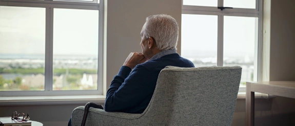 Alter Mann sitzt alleine in einem Sessel und starrt durch das Fenster in die Stadt.