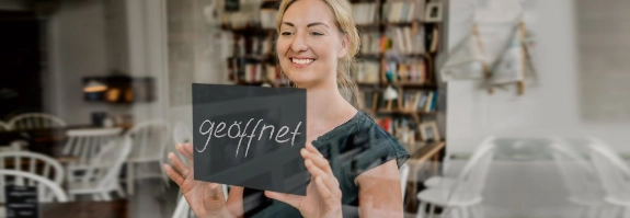 Frau hält Tafel mit "geoffnet" Schriftzug in die Kamera, lächelt, Bücherschrank im Hintergrund