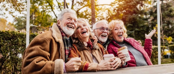 Vier ältere Menschen lachen zusammen im Freien, herbstliche Umgebung mit grünen Hecken im Hintergrund.