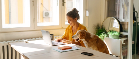 Frau mit Hund arbeitet am Laptop am Schreibtisch, helles Büro mit Fenster und Pflanzen im Hintergrund.