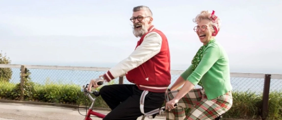 Älteres Paar auf rotem Tandem-Fahrrad an Uferpromenade, lachend, Wasser und Grün im Hintergrund