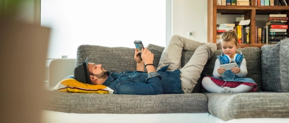 Zwei Kinder liegen auf grauem Sofa, lesen Bücher, während ein Erwachsener zwischen ihnen sitzt. Gemütliches Wohnzimmer mit Bücherregal im Hintergrund.