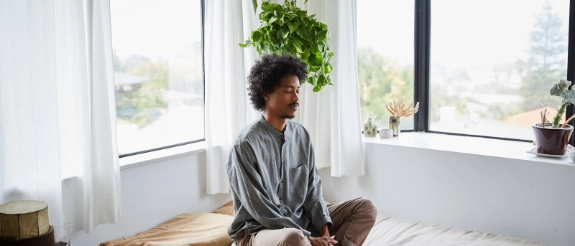 Person sitzt meditierend auf Bett am Fenster mit Blick nach außen, helles Schlafzimmer mit Pflanzen