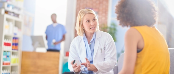 Ärztin in weißem Kittel spricht mit Patientin in gelbem Top in einer medizinischen Klinik