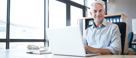 Älterer Mann mit grauem Haar arbeitet konzentriert am Laptop an seinem Schreibtisch im hellen Büro.