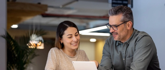 Zwei Personen schauen gemeinsam auf einen Tablet-Bildschirm in einem modernen Büro mit Deckenleuchten und Pflanze.