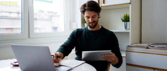 Mann sitzt am Schreibtisch in einer Küche, arbeitet am Laptop, trägt Kopfhörer, helles Loft-Büro mit Fenster