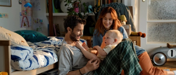 Familie spielt mit Baby im Kinderzimmer, umgeben von Spielzeug und Büchern auf dem Boden.