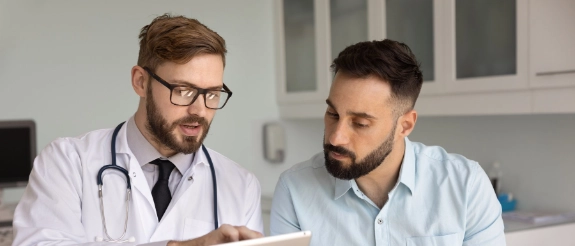 Arzt in Kittel und Brille bespricht mit Patient Befunde auf Tablet in Klinik
