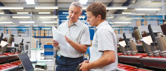 Zwei Männer in Industriehalle besprechen Dokument vor Werkzeugregalen und Maschinen