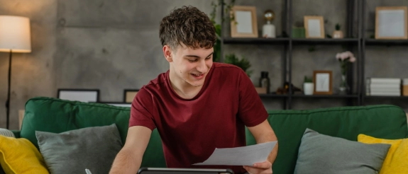 Junger Mann in rotem Shirt sitzt auf grünem Sofa und arbeitet konzentriert an Tablet, modernes Wohnzimmer im Hintergrund