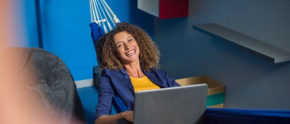Frau mit lockigem Haar in blauem Blazer und gelbem Top arbeitet am Laptop in modernem Büro mit blauer Wand
