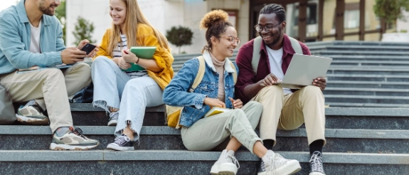 Vier junge Menschen sitzen auf Treppen und nutzen zusammen Smartphones und Laptops im Freien.