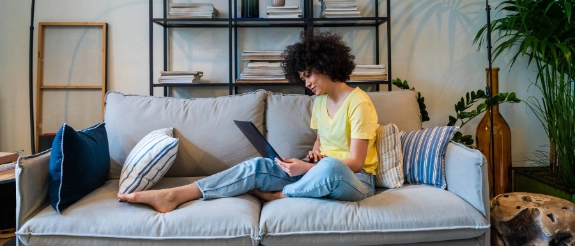 Frau mit lockigem Haar sitzt auf Sofa in gelbem Shirt und arbeitet am Laptop, umgeben von Kissen und Pflanzen im modernen Wohnzimmer.