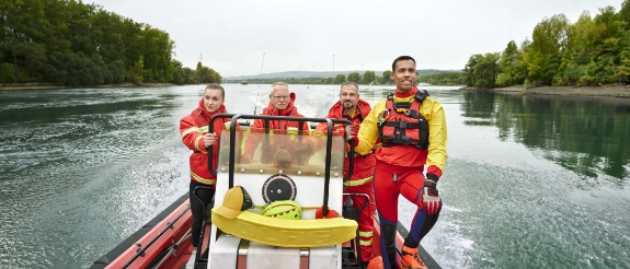 Vier Personen in Rettungswesten auf einem Rettungsboot auf einem Fluss mit Landschaft im Hintergrund