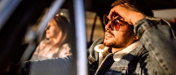 Mann mit Sonnenbrille sitzt im Auto, blickt konzentriert nach vorne, grüne Jacke, Innenraum des Fahrzeugs sichtbar.