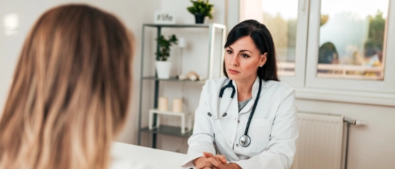 Ärztin im weißen Kittel mit Stethoskop führt Patientengespräch am Schreibtisch in moderner Arztpraxis.