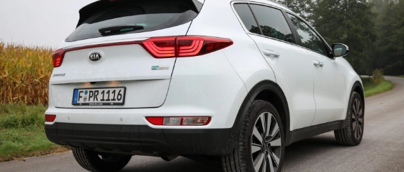 Weißes SUV-Fahrzeug mit roten Rückleuchten von hinten, geparkt auf Landstraße mit Grünflächen und Bäumen im Hintergrund