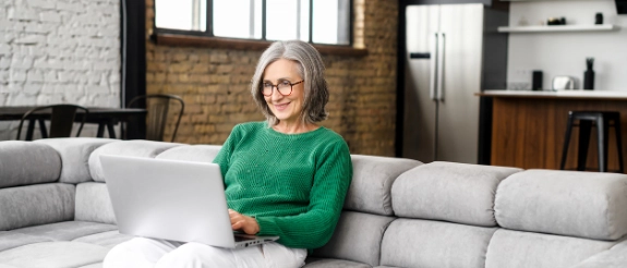 Frau mit grauem Haar und Brille arbeitet am Laptop auf grauem Sofa im modernen Wohnzimmer mit Ziegelmauer.