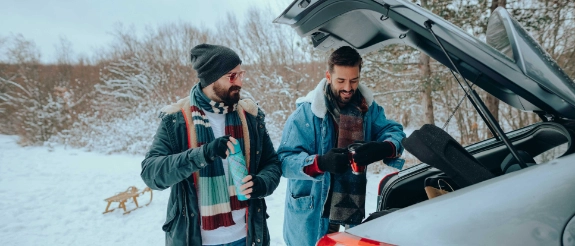 Zwei Personen öffnen Kofferraum eines Autos in verschneiter Winterlandschaft mit Schneekoffer und Ausrüstung.