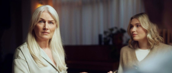Blonde Frau in hellem Blazer sitzt am Tisch mit Teller, ernsthafte Mimik, warme Beleuchtung im Hintergrund