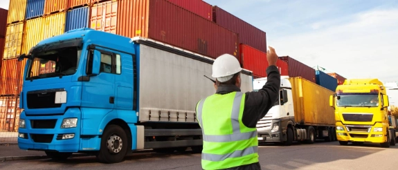 Arbeiter in Warnweste und Schutzhelm dirigiert Lastkraftwagen auf Hafengelände mit gestapelten Containern.