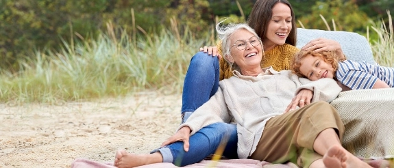Drei Generationen Frauen sitzen lachend zusammen am Strand, umgeben von Strandhafer und grüner Natur.